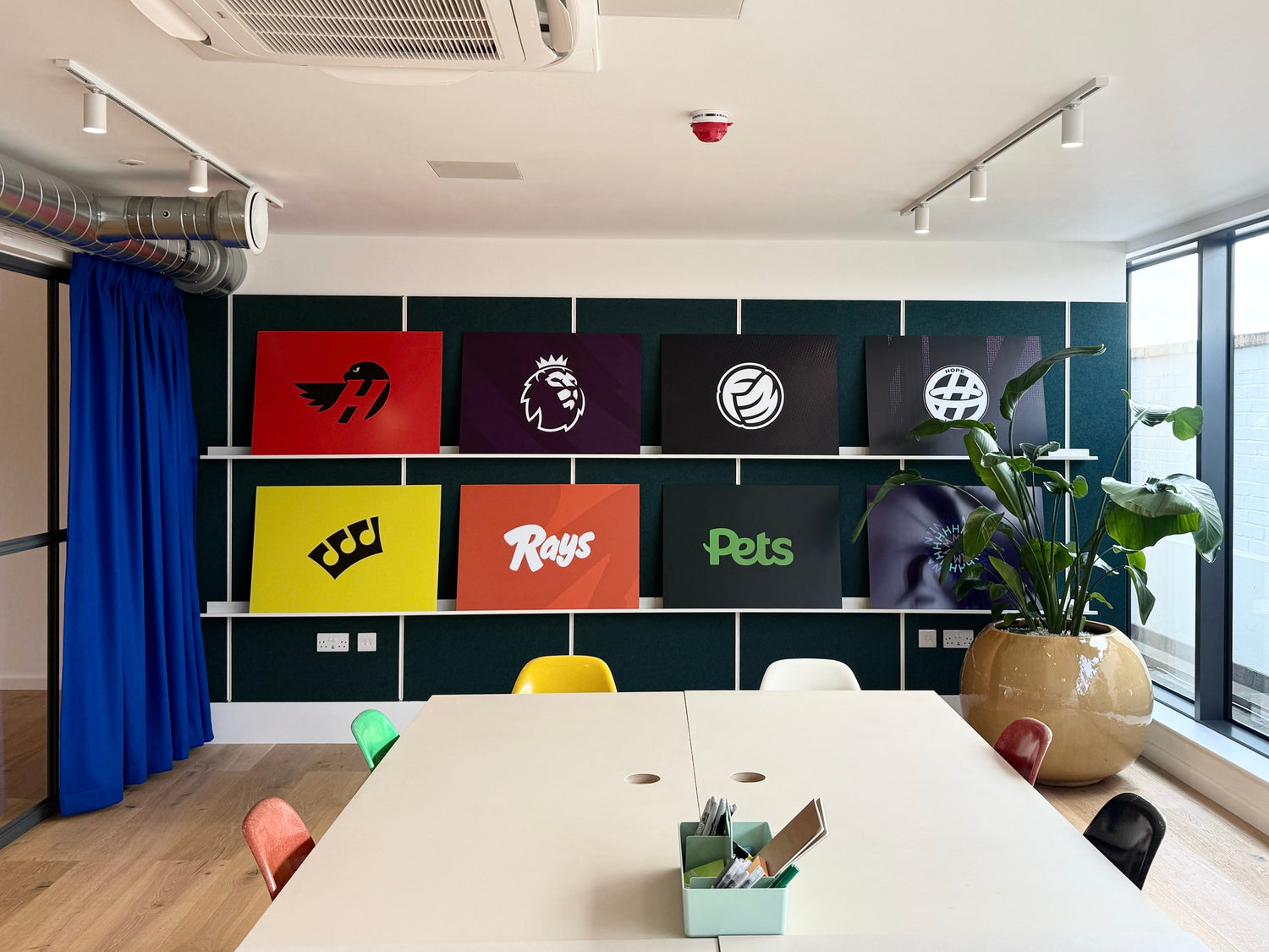 A modern office or meeting room interior with a long white table in the foreground, track lighting, wood flooring, a blue curtain, and a dark wall displaying colorful sports/team logos on white ON&ON mini shelves.