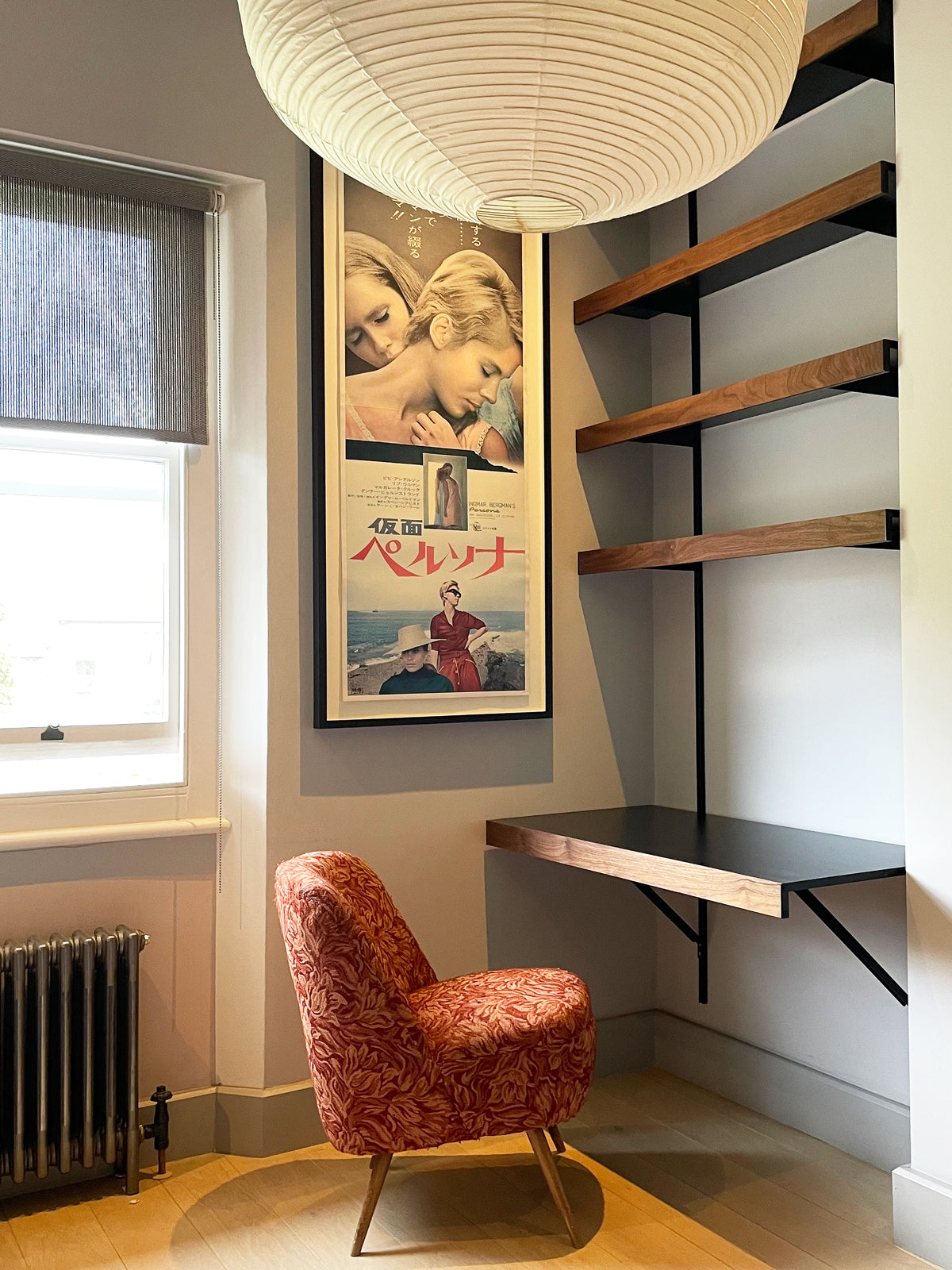 A luxury styled interior corner featuring a vintage Japanese movie poster, a red patterned armchair, a floating desk with walnut shelves above, and a large white paper lamp shade hanging from the ceiling.