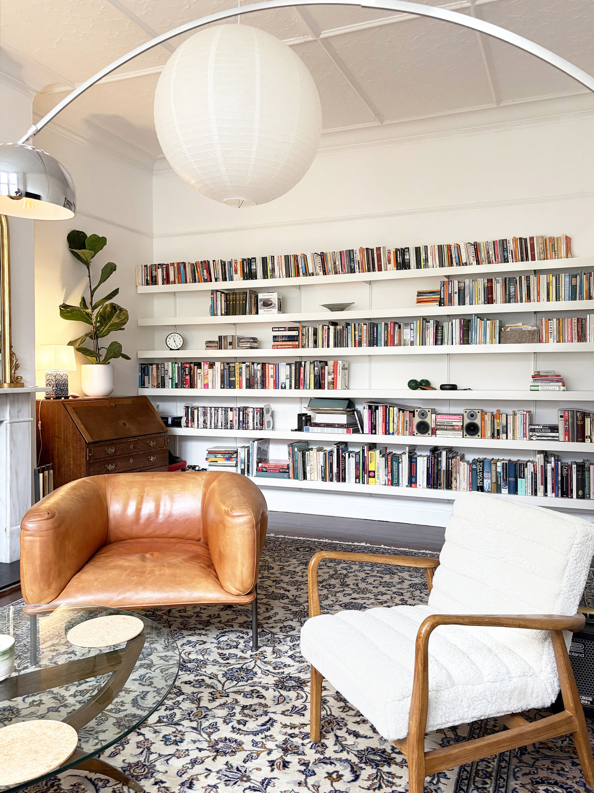 A cozy, well-lit living room featuring large white bookshelves filled with books, a brown leather armchair, a white upholstered wooden armchair, a patterned rug, and a white spherical pendant light hanging from an arched floor lamp.