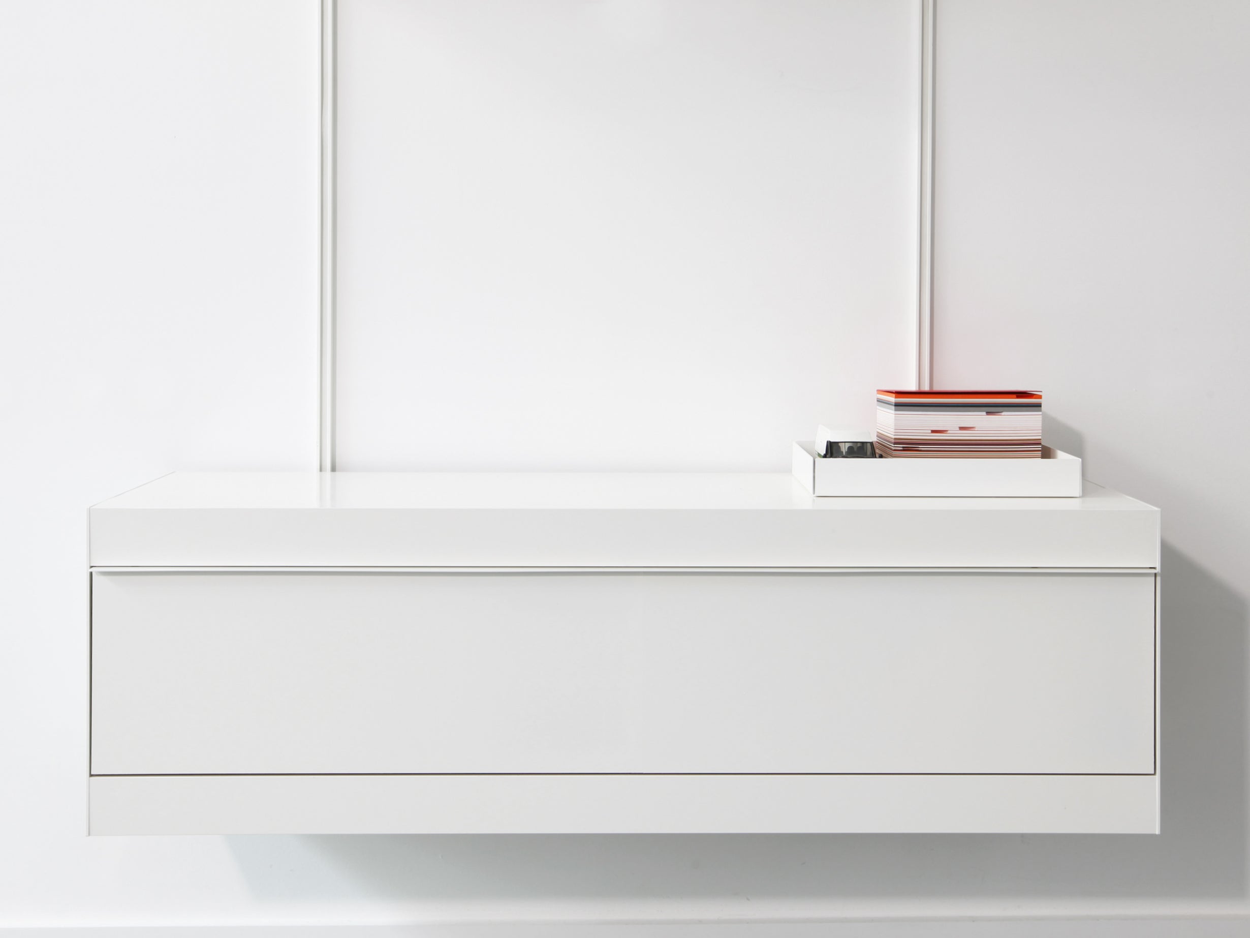 A minimalist white wall-mounted cabinet with a small stack of books and a tray on top, set against a clean white wall.