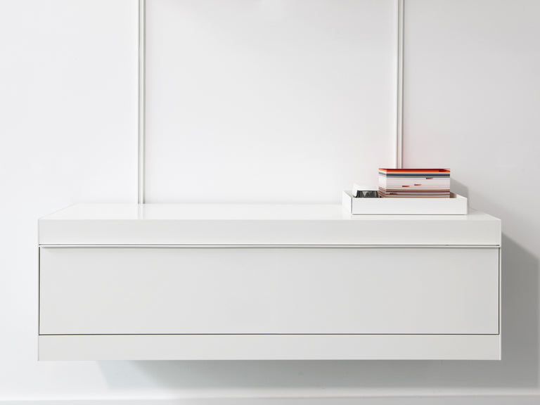 A minimalist white wall-mounted cabinet with a small stack of books and a tray on top, set against a clean white wall.