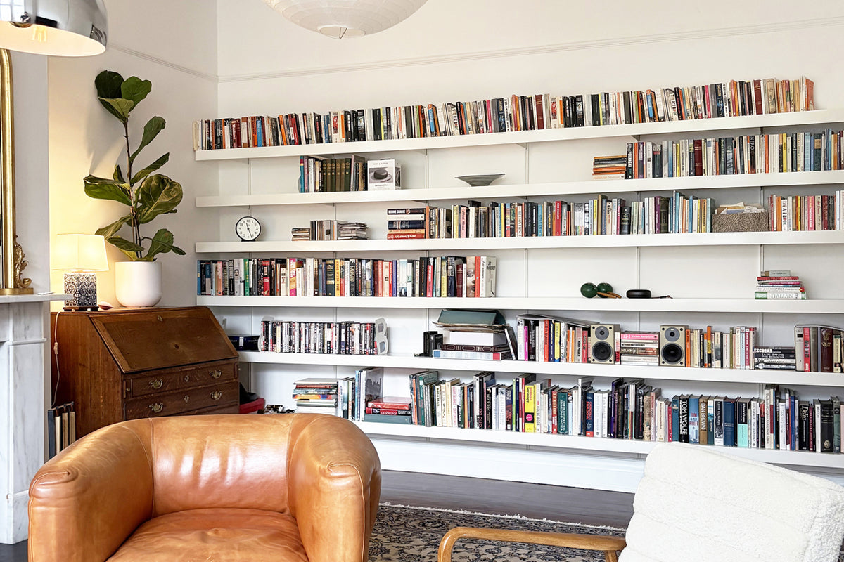 A large white built-in bookshelf system mounted on a white wall in a bright room, filled with books and small decor items. A tan leather armchair is in the foreground, along with a potted plant, a wooden desk, and part of another chair.