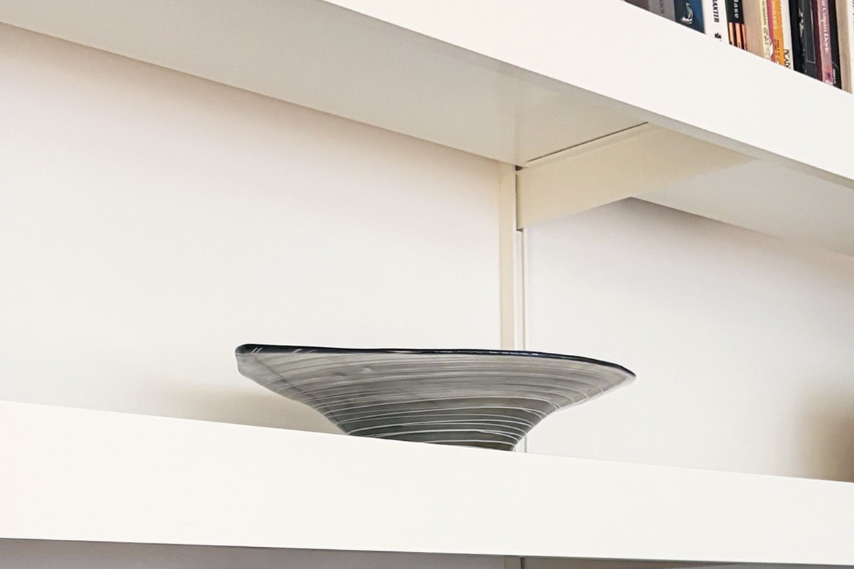 A modern, shallow conical bowl with a textured ribbed surface and a dark rim, displayed on a minimalist white bookshelf showing shelving fixings and bracket.