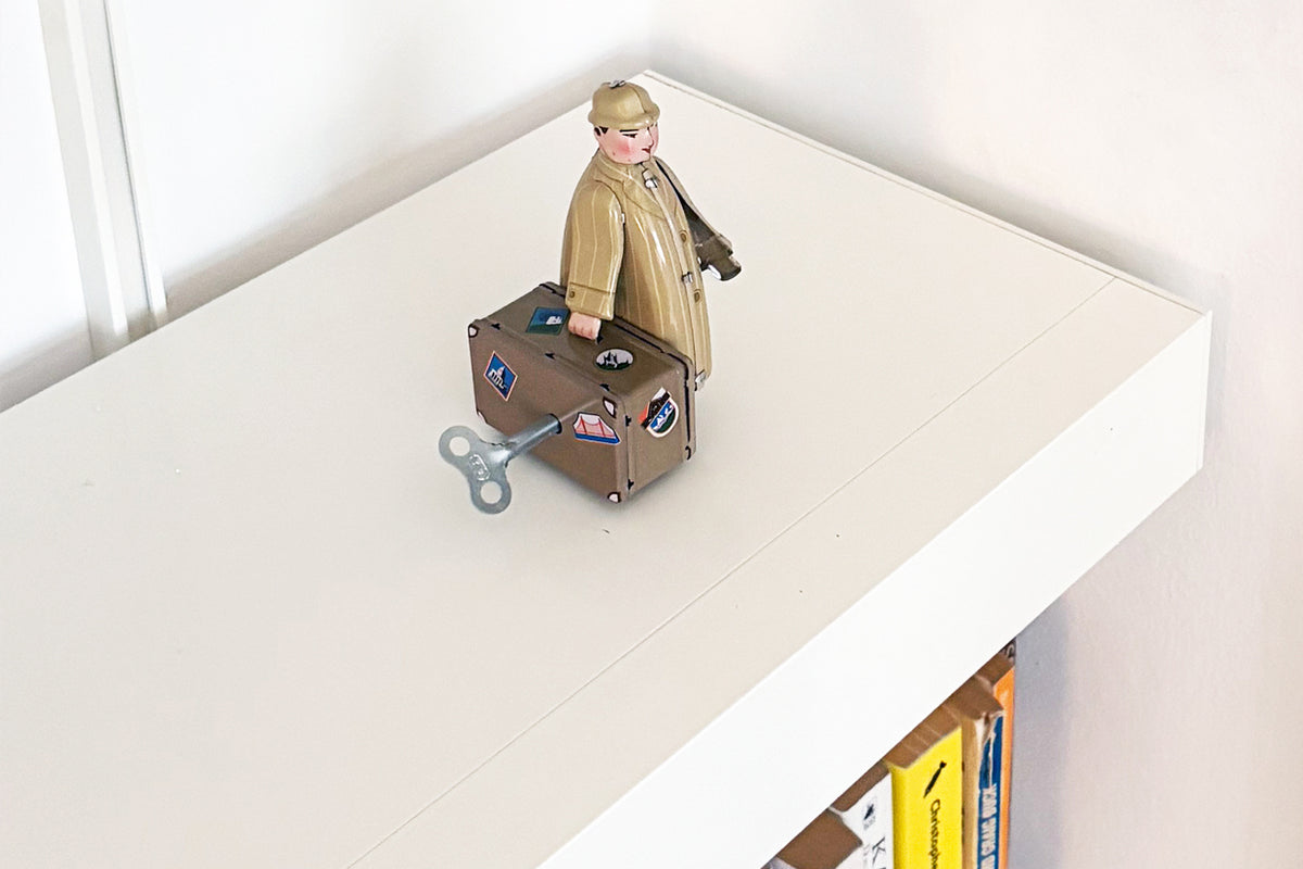 A vintage-style wind-up toy figure of a man in a tan coat standing next to a brown suitcase with stickers, set on a white aluminium shelf.