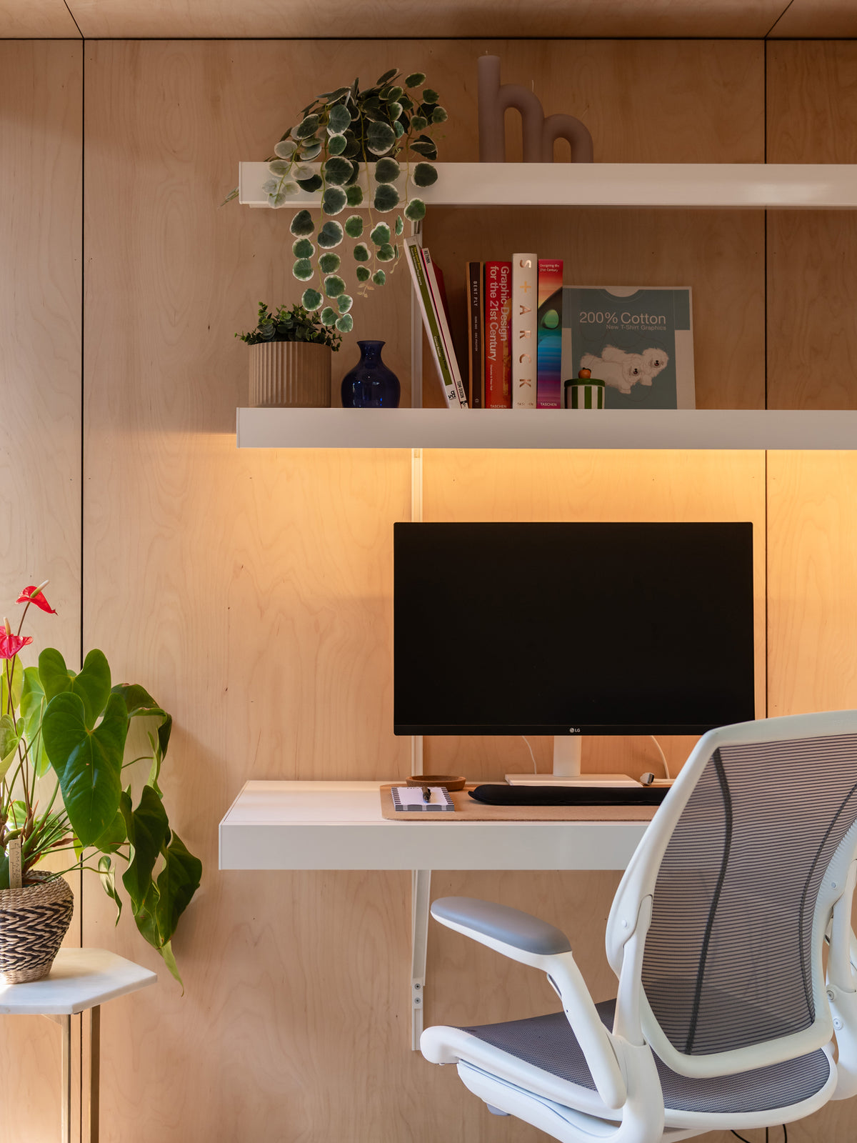 A modern home office setup featuring a white floating desk, a black computer monitor, a white ergonomic mesh office chair, and a light wood paneled wall with built-in shelves holding books, a plant, and decorative items.