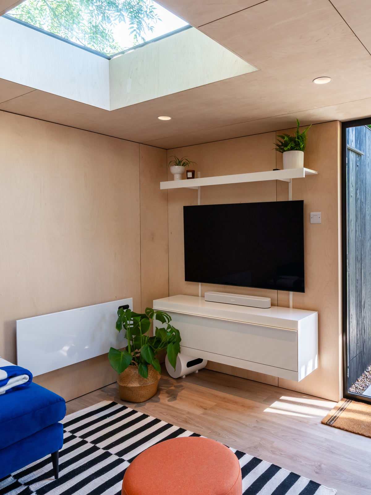 A modern living room corner featuring a wall-mounted black flat-screen TV above a white floating media console system and shelf, with a black-and-white striped rug, orange pouf, blue chair, and potted green plants in a light wood interior.