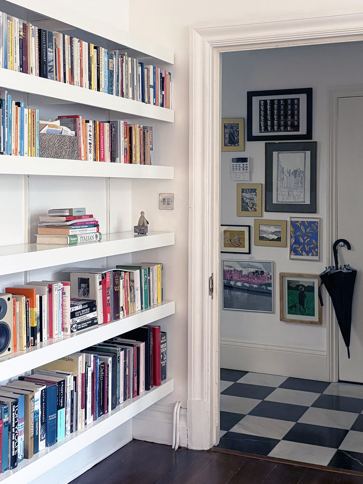 White floating bookshelves filled with books in a home interior, with a doorway leading to a hallway featuring framed artwork, a black-and-white checkered floor, and a closed black umbrella hanging on the door.