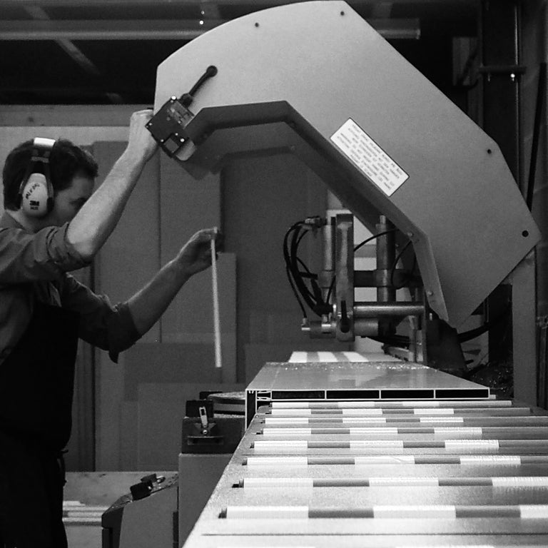 A black and white photograph showing a worker wearing ear protection and adjusting a large metal machine cutting aluminium shelves.