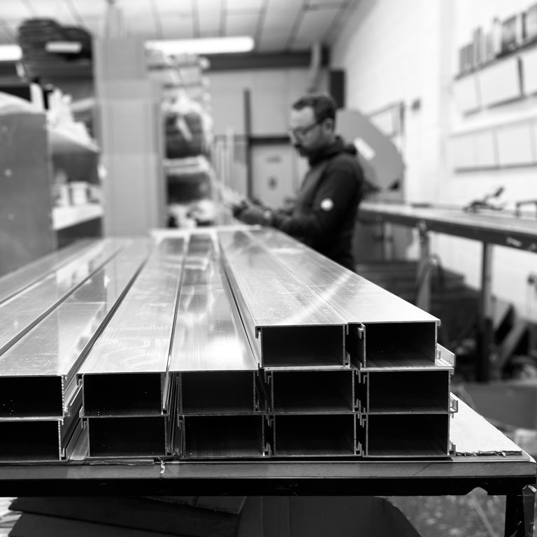 A black and white photograph showing a stack of long, aluminium channel profiles on a workbench in a workshop, with a blurred worker visible in the background.