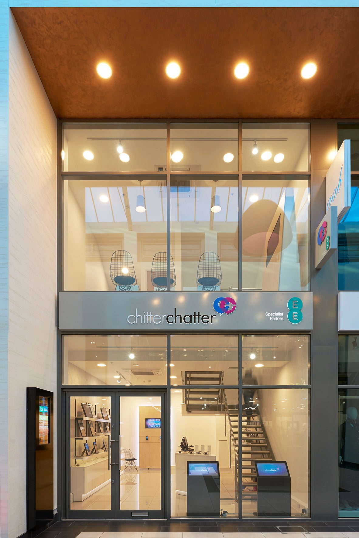 The exterior of the Chitter Chatter retail store, featuring a modern glass facade, a two-story layout, a warm brown textured ceiling, and illuminated interior spaces.