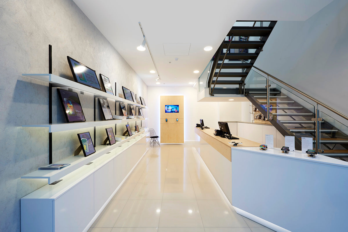 A retail phone shop with a long white reception desk, a wall-mounted display of multiple monitors and phones, a light wood door at the end, and a black staircase on the right.